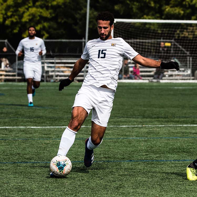 Soccer player in a white uniform with number 15 dribbles a ball on a grassy field during a daytime match. Another player is visible in the background, and a goal post is seen behind them.