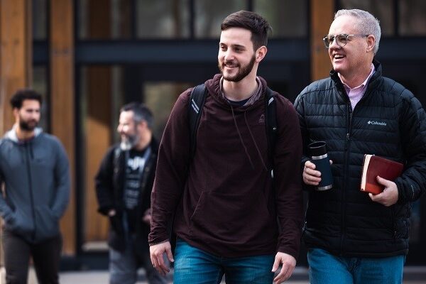 A younger man with a backpack and an older man holding a coffee cup and a book walk together outside, smiling. Two other men in casual clothing are in the background, also smiling in a lively outdoor setting.