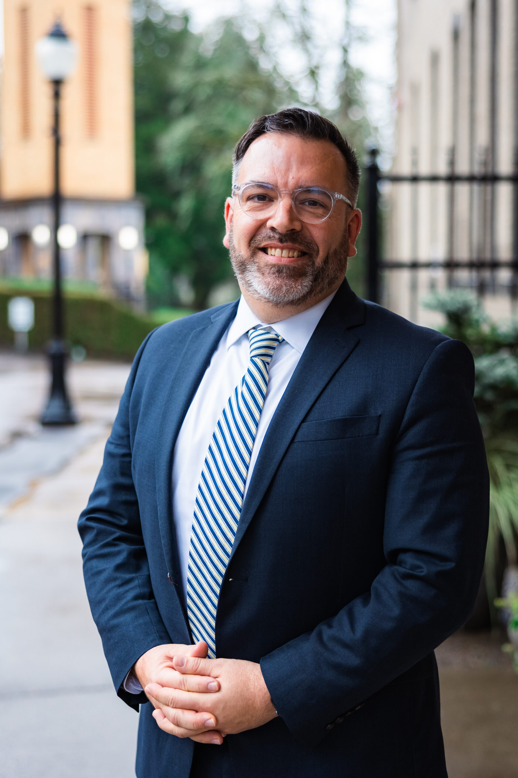 A man in a blue suit and striped tie smiles confidently with hands clasped in front of him. He stands on a wet sidewalk, with blurred buildings and greenery in the background.