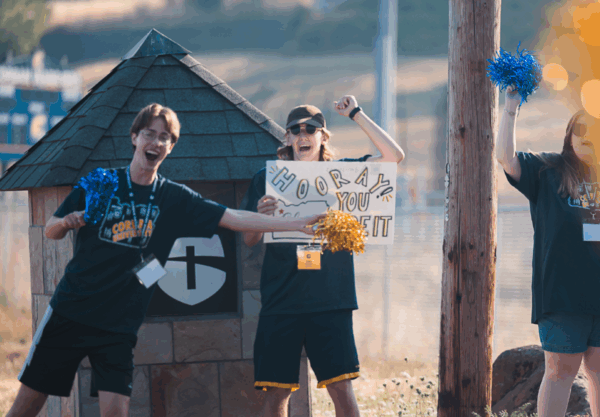Three people cheer with pom-poms and a sign saying Hooray You Did It in a sunny outdoor setting. They wear casual shirts and name tags, with a small stone structure and sports field in the background.
