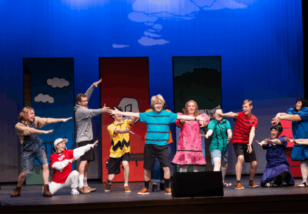 A theater group performs on stage, with actors dressed as colorful, cartoon-like characters. They strike dramatic poses in front of a backdrop featuring simple buildings and trees. A yellow booth with Psychiatric Help 5¢ is on the left.