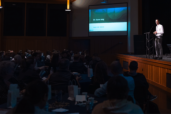 A speaker stands on a stage in front of a seated audience, giving a presentation. A screen displays Dr. Aaron Minj and a title. The room is dimly lit, with focus on the speaker and the screen.