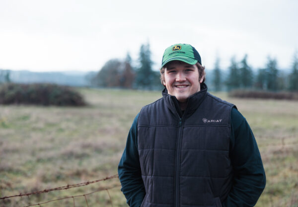 A person wearing a green cap and dark vest smiles while standing in a grassy field with trees in the background. The scene appears outdoors and cloudy.