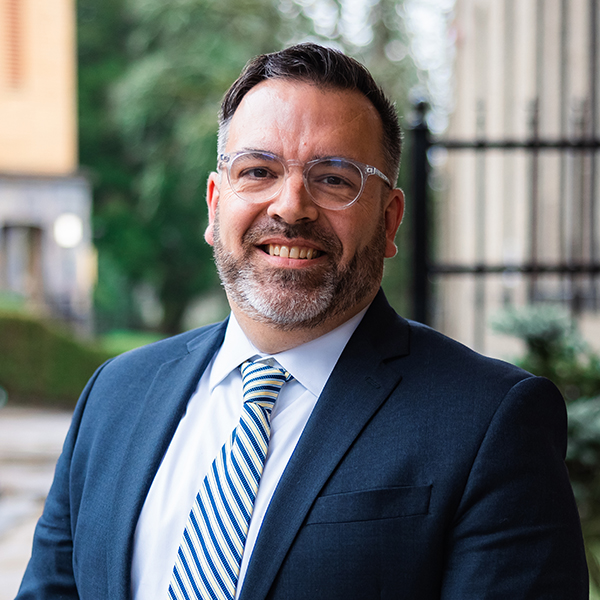 Portrait of Dr. John Mark Yeats, President of Corban University. The background features greenery and building elements.