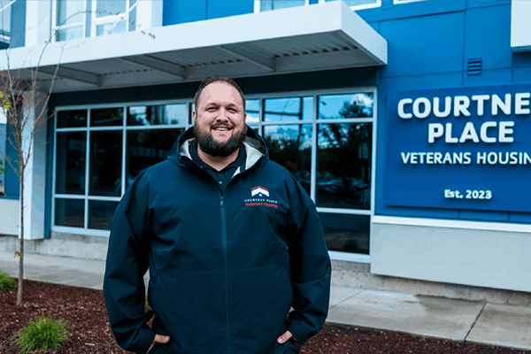 A person stands in front of a building with a sign that reads Courtney Place Veterans Housing, Est. 2023. They are wearing a dark jacket with a white and red logo. The entrance is visible in the background, and some small plants are in front.