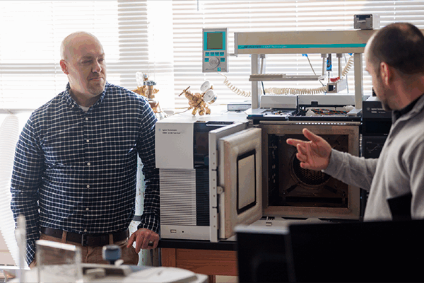 Two men are in a laboratory with a large machine that has an open compartment. One is explaining something while gesturing towards the machine. They are both wearing casual clothing. Sunlight streams through the window blinds behind them.