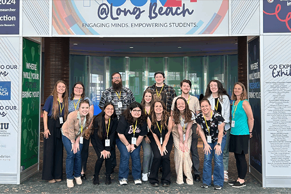 A group of 14 people stands smiling in front of a conference entrance with signage reading Engaging Minds. Empowering Students. They are inside a building with glass doors and various banners around them.