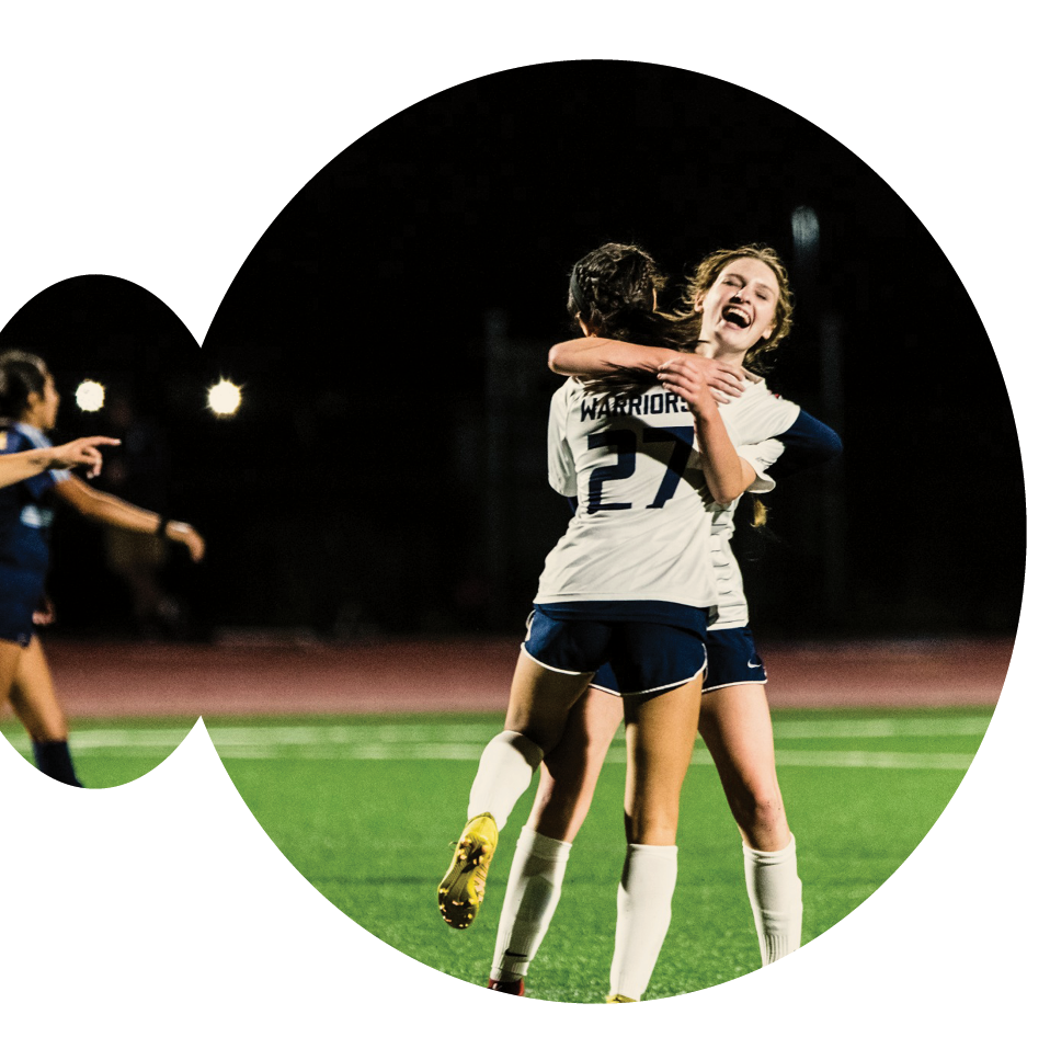 Two soccer players in white jerseys, from a Christian college in Oregon, are joyfully embracing on a dimly lit field at night, celebrating. One proudly wears the number 27. In the backdrop, another player is partly visible on the left.