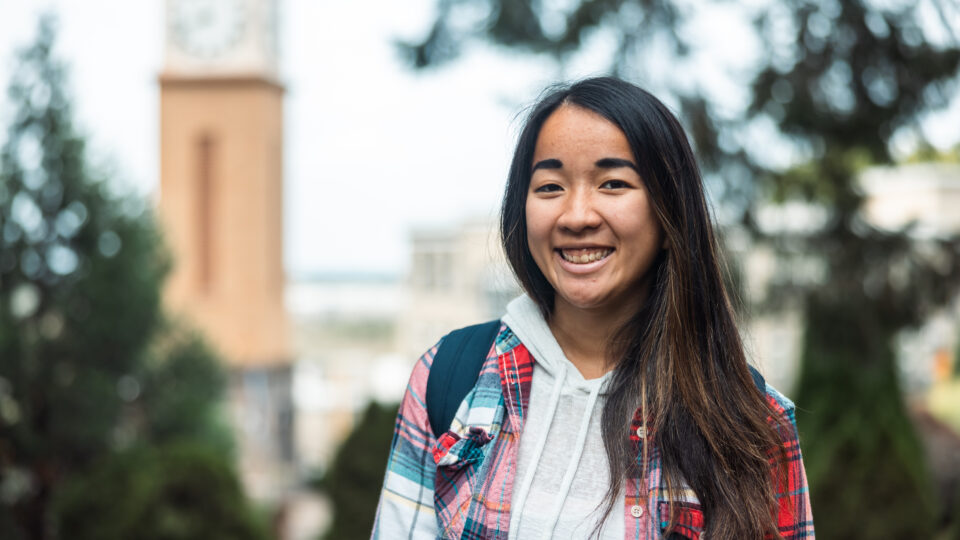 A person with long hair smiles while wearing a plaid shirt over a hoodie and carrying a backpack. They stand in front of a blurred outdoor background with trees and a clock tower.