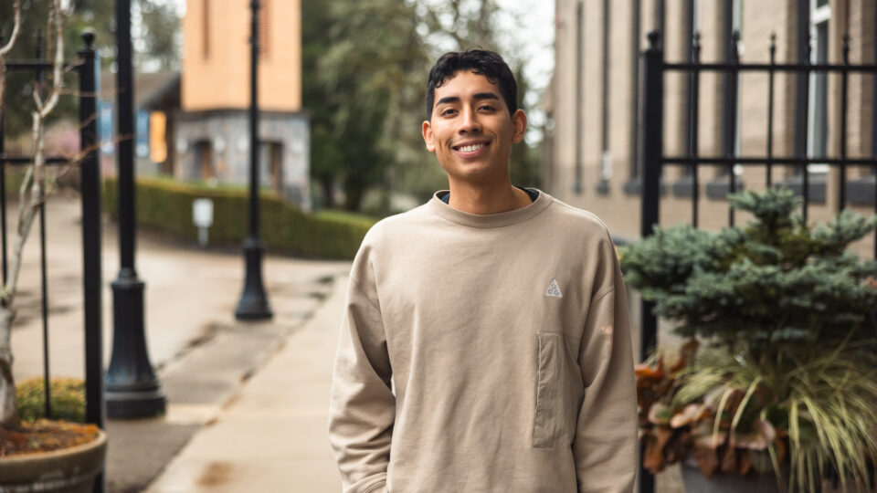 A person wearing a beige sweatshirt smiles while standing on a sidewalk with potted plants and black railings. The background features a building and trees. The scene is overcast, suggesting a cool day.