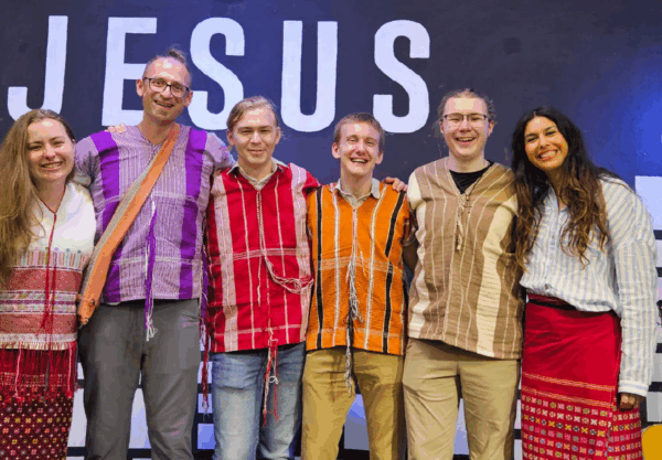A group of six smiling people stand side by side, wearing colorful traditional outfits. They are positioned in front of a wall with the word JESUS displayed prominently at the top. A guitar is partially visible on the right side.