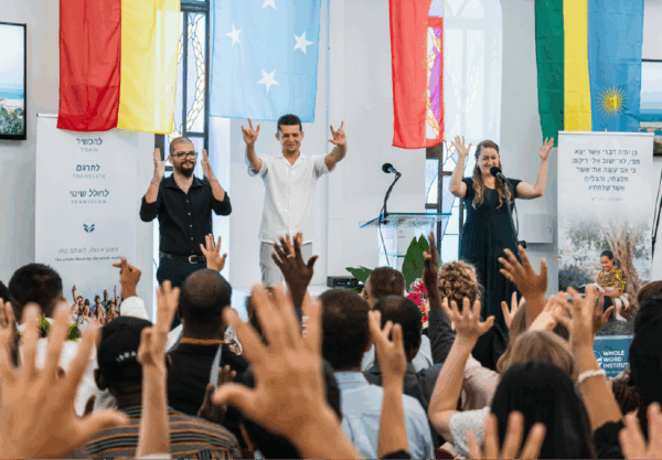 Three people stand on stage, smiling and gesturing with peace signs, facing an audience with raised hands. Behind them are colorful flags and posters. The atmosphere is lively and engaging.