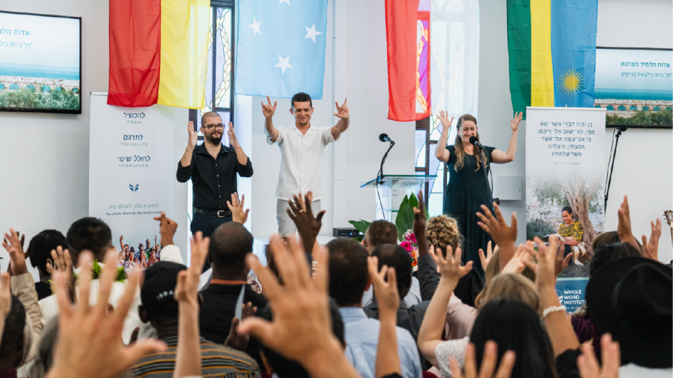 Three people stand on stage, smiling and gesturing with peace signs, facing an audience with raised hands. Behind them are colorful flags and posters. The atmosphere is lively and engaging.