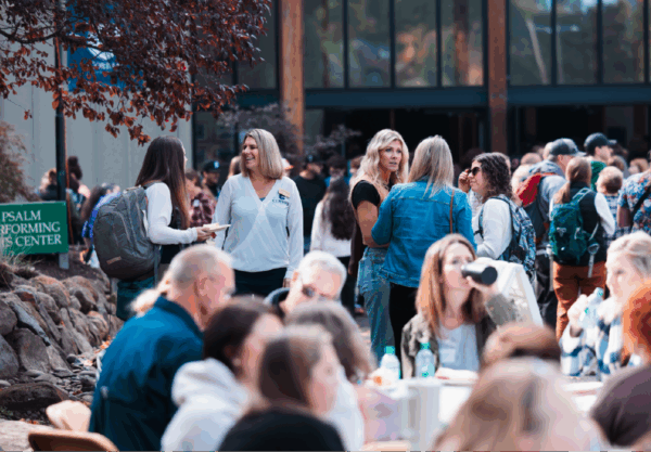 A group of people gathered outdoors at an event near a building with a sign that reads Salem Performing Arts Center. Some are seated at tables, while others stand and chat. Trees and rocks are part of the landscape.