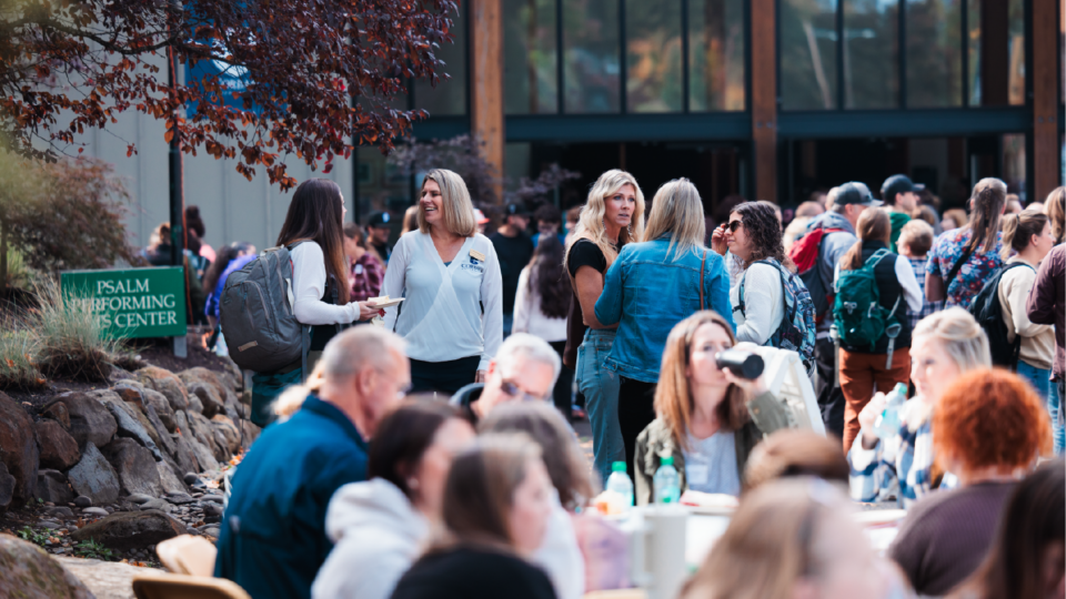 A group of people gathered outdoors at an event near a building with a sign that reads Salem Performing Arts Center. Some are seated at tables, while others stand and chat. Trees and rocks are part of the landscape.