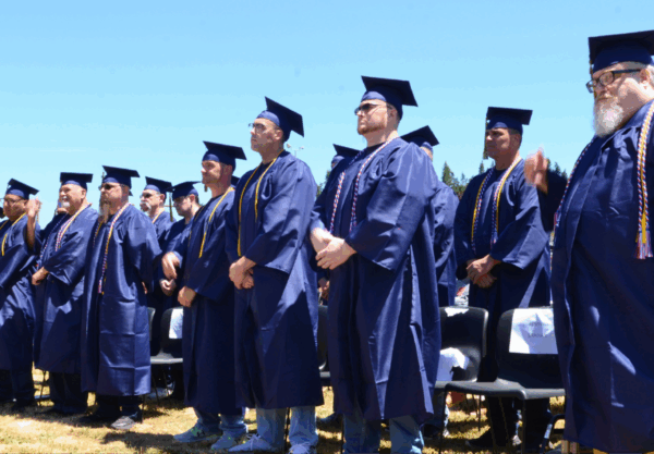 A group of graduates in navy blue caps and gowns stand together outdoors on a sunny day. They wear tassels and cords, and some are adjusting their attire. Chairs and a clear blue sky are visible in the background.