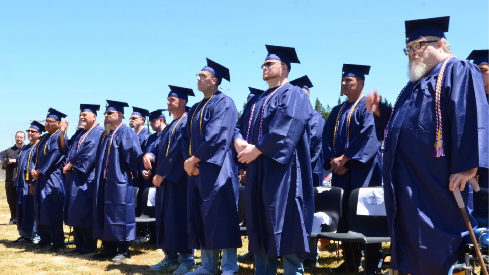 A group of graduates in navy blue caps and gowns stand together outdoors on a sunny day. They wear tassels and cords, and some are adjusting their attire. Chairs and a clear blue sky are visible in the background.