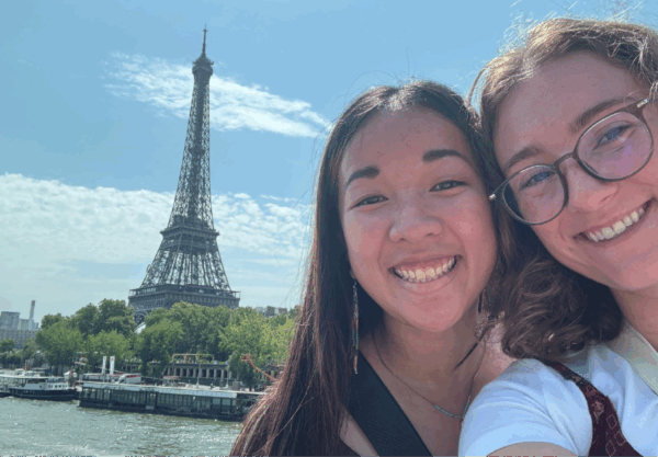 Two smiling individuals pose for a selfie in front of the Eiffel Tower on a sunny day. The river and trees are visible in the background, adding a vibrant touch to the iconic Parisian scene.
