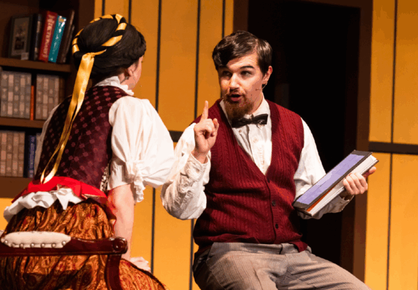 A man in a red vest and bow tie holds a book while gesturing with his finger, sitting across from a woman in a historical costume with a yellow ribbon in her hair. They appear to be in a library with bookshelves in the background.