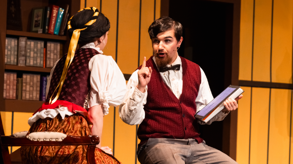A man in a red vest and bow tie holds a book while gesturing with his finger, sitting across from a woman in a historical costume with a yellow ribbon in her hair. They appear to be in a library with bookshelves in the background.