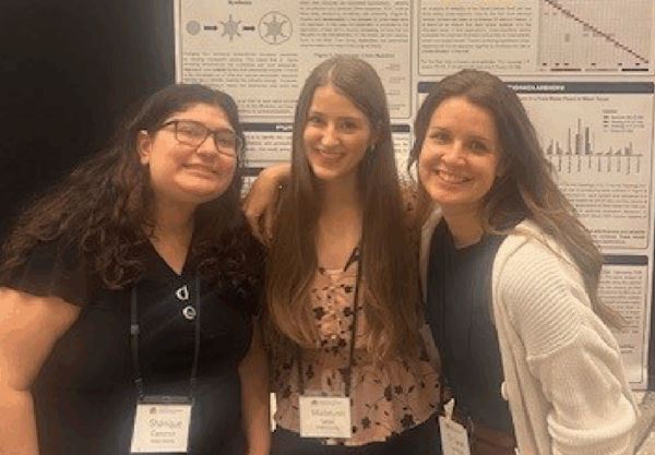Three people stand together, smiling, in front of a scientific poster. They are wearing name tags and appear to be at a conference or presentation.