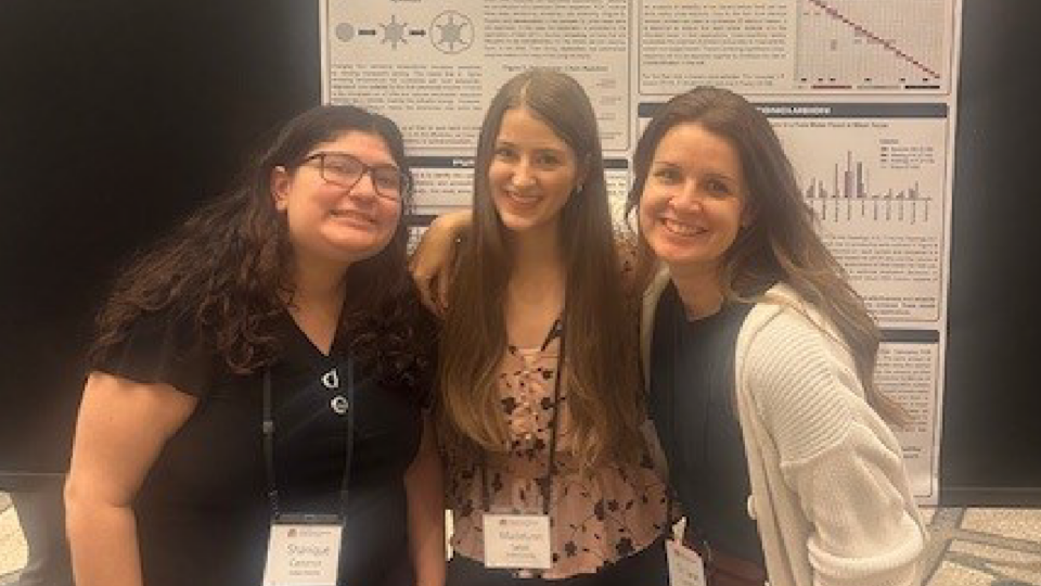 Three people stand together, smiling, in front of a scientific poster. They are wearing name tags and appear to be at a conference or presentation.