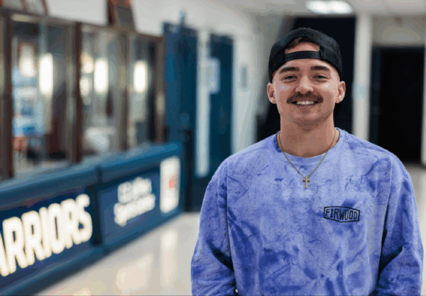 A person wearing a purple tie-dye sweatshirt and a backward black cap stands smiling in a hallway. Theres a sign reading Warriors in the background. The person has a mustache and wears a necklace with a cross pendant.