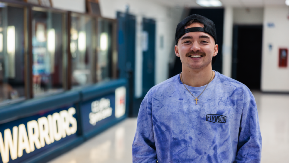 A person wearing a purple tie-dye sweatshirt and a backward black cap stands smiling in a hallway. Theres a sign reading Warriors in the background. The person has a mustache and wears a necklace with a cross pendant.