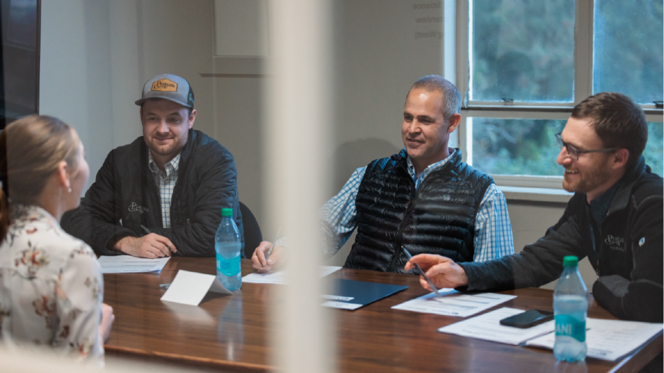 A woman sits at a table during a meeting with three men. Two men wear vests and one wears a cap. Papers and water bottles are on the wooden table. A window with trees visible is in the background.
