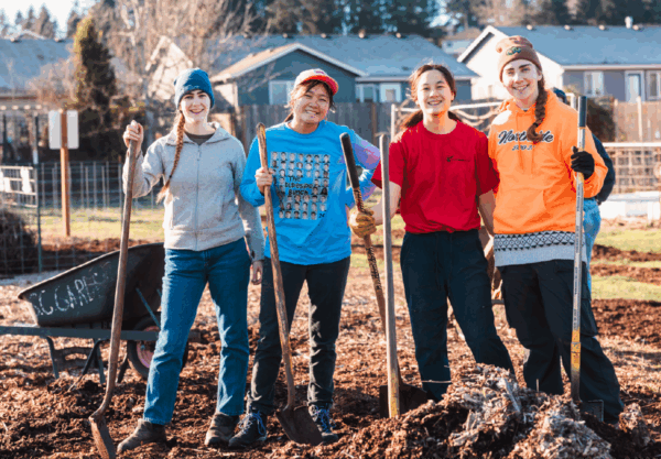 Four people smiling and holding shovels stand on a dirt patch in a community garden. They are dressed in casual outdoor clothing, with gloves and hats. In the background, theres a fence, buildings, and a wheelbarrow filled with tools.