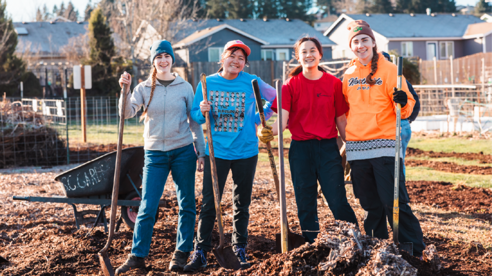 Four people smiling and holding shovels stand on a dirt patch in a community garden. They are dressed in casual outdoor clothing, with gloves and hats. In the background, theres a fence, buildings, and a wheelbarrow filled with tools.
