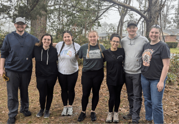 Seven people standing in a row outside, smiling. They are dressed casually, wearing sneakers and comfortable clothing. Trees and a house are visible in the background. The group appears to be enjoying a day outdoors.