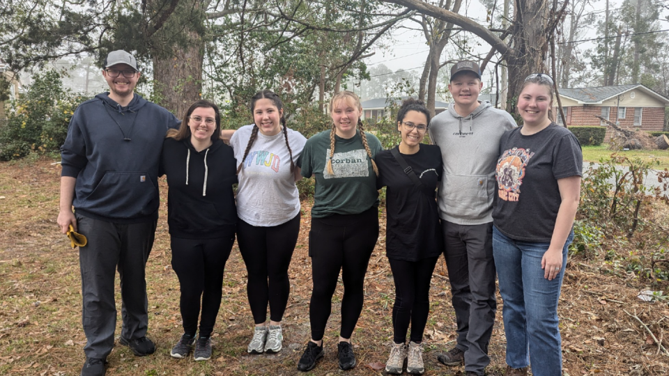 Seven people standing in a row outside, smiling. They are dressed casually, wearing sneakers and comfortable clothing. Trees and a house are visible in the background. The group appears to be enjoying a day outdoors.