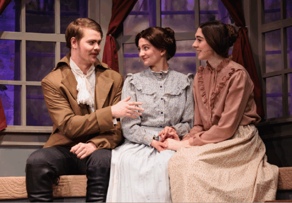 Three actors in period costumes sit on a window seat with red curtains. The man on the left gestures while the two women on the right listen intently, creating a scene suggestive of a theatrical performance or historical drama.