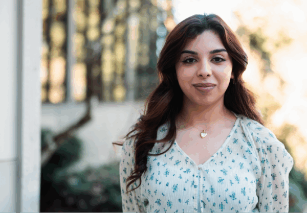 A woman with long, dark hair stands outside, wearing a light-patterned blouse and a heart-shaped necklace. She is smiling slightly with trees and a building in the blurred background.