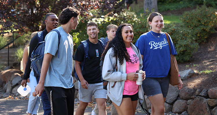 A group of six college students, smiling and talking, walk outdoors along a path lined with greenery. Some carry backpacks and drinks, suggesting they are on a campus.