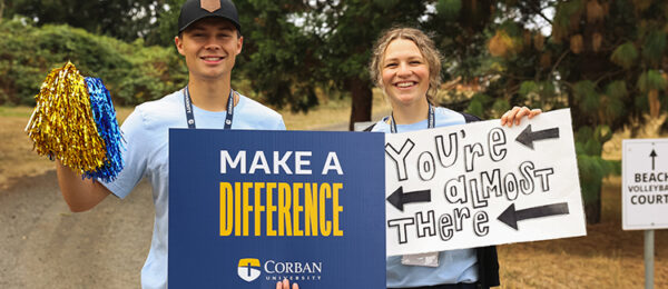 Two smiling people stand outdoors. One holds gold and blue pom-poms and a sign that reads “MAKE A DIFFERENCE, CORBAN UNIVERSITY.” The other holds a hand-drawn sign that says “You’re almost there” with arrows. Trees are in the background.