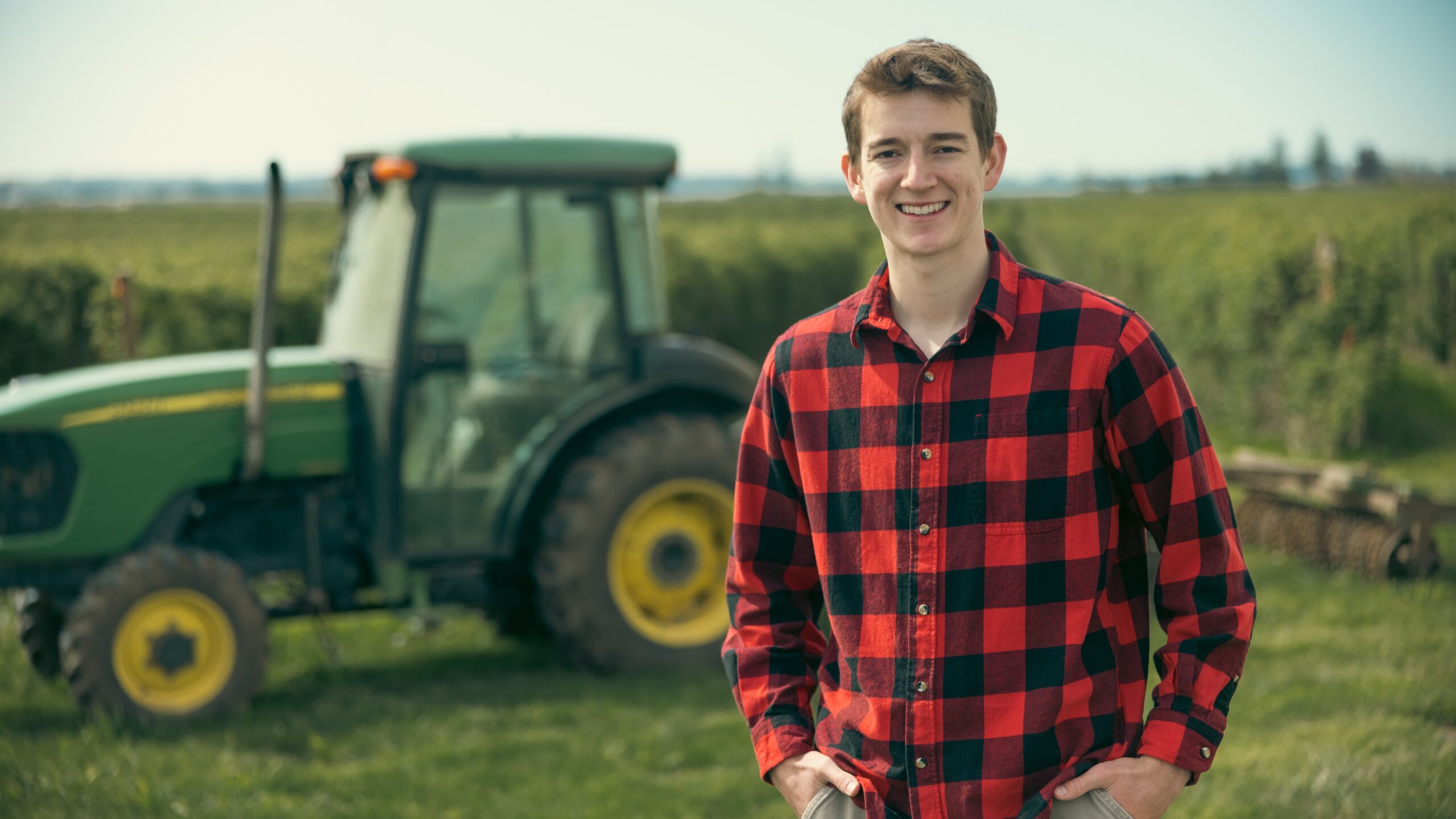 A young man wearing a plaid shirt poses in front of a large tractor, showcasing a rural farming setting.
