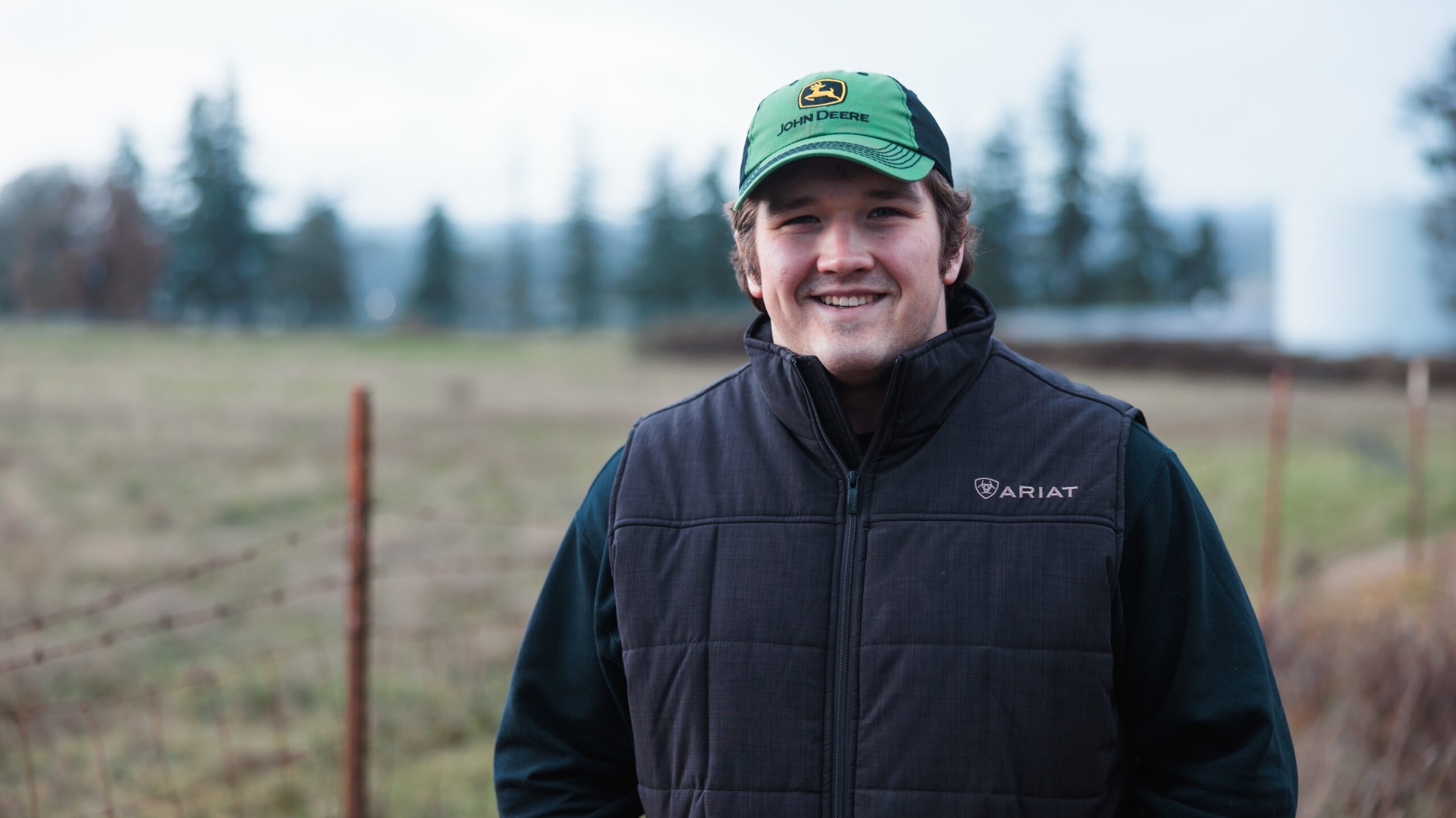 A young man in a green hat and vest stands in a field, smiling and enjoying the outdoors.