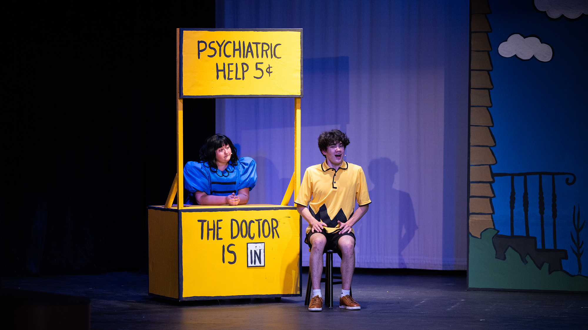 Actors portraying Charlie Brown and Lucy sitting on a stage, performing a scene from the musical "You're a Good Man, Charlie Brown."