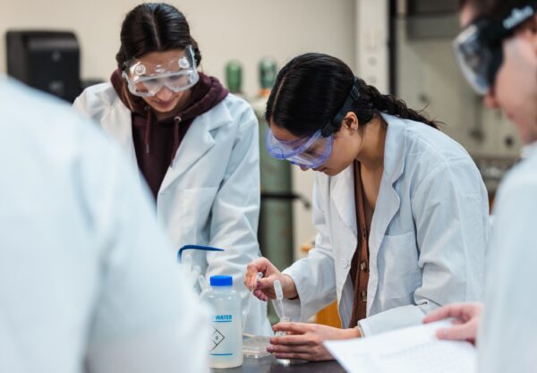 A group of students wearing lab coats and goggles engage in hands-on experimentation in a lab environment.