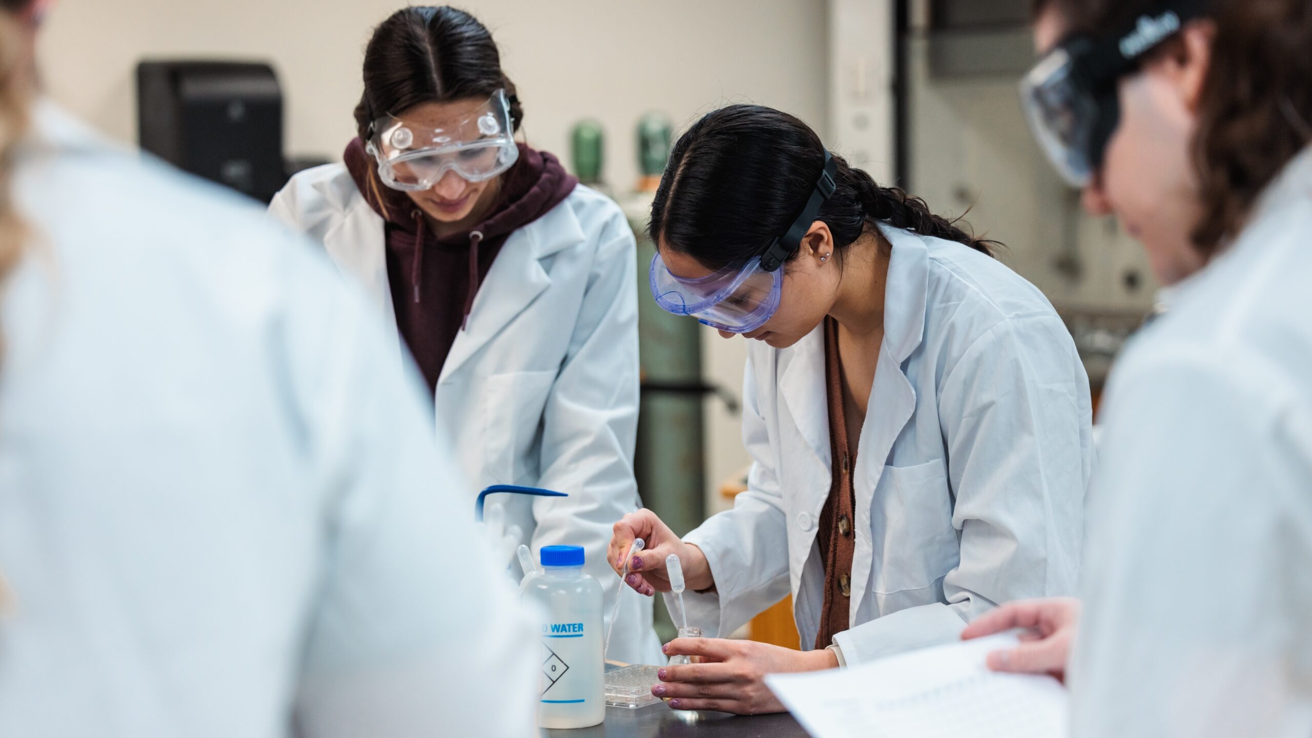 A group of students wearing lab coats and goggles engage in hands-on experimentation in a lab environment.