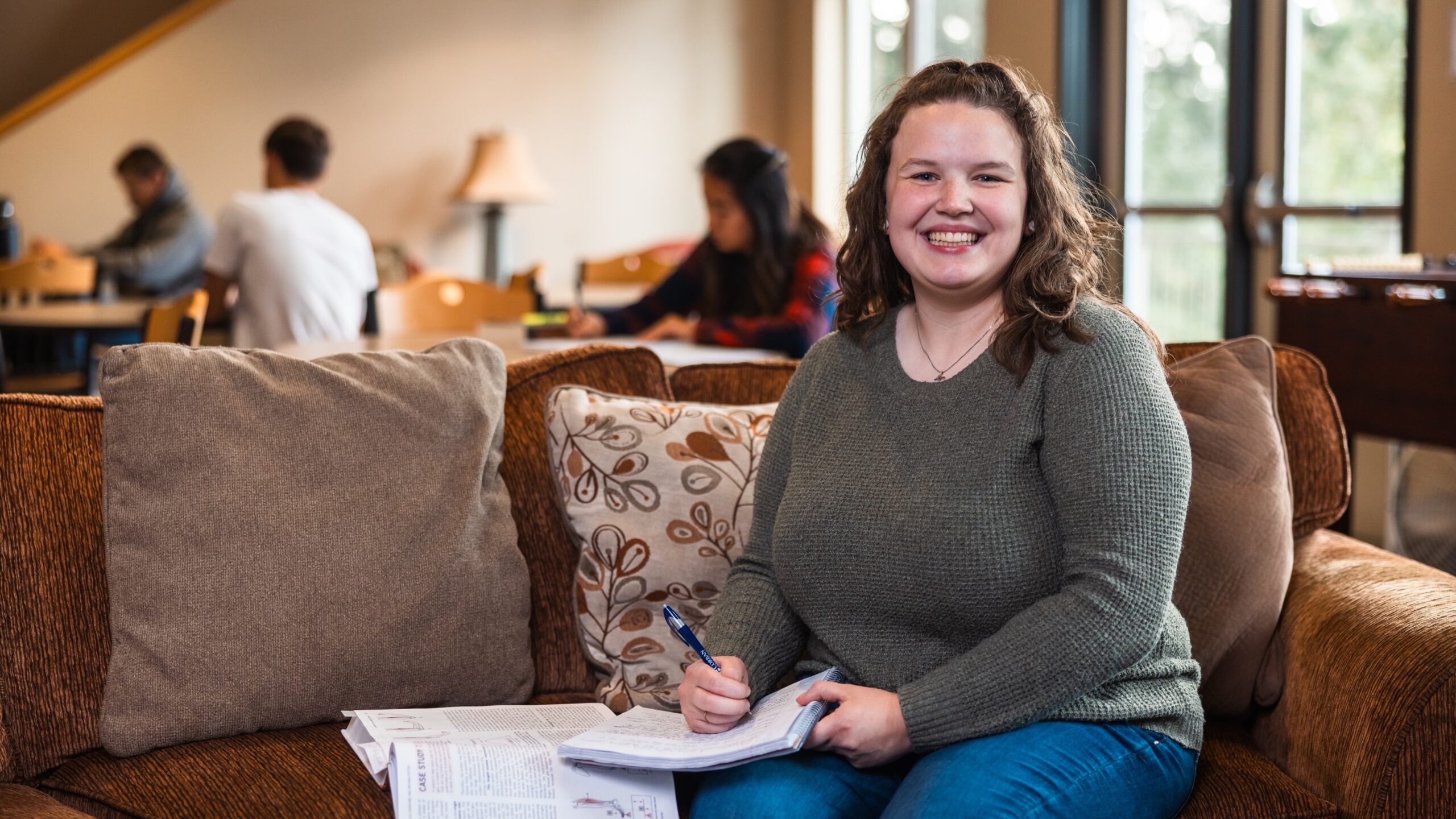A smiling woman sits on a couch, holding a pen and paper, engaged in writing.
