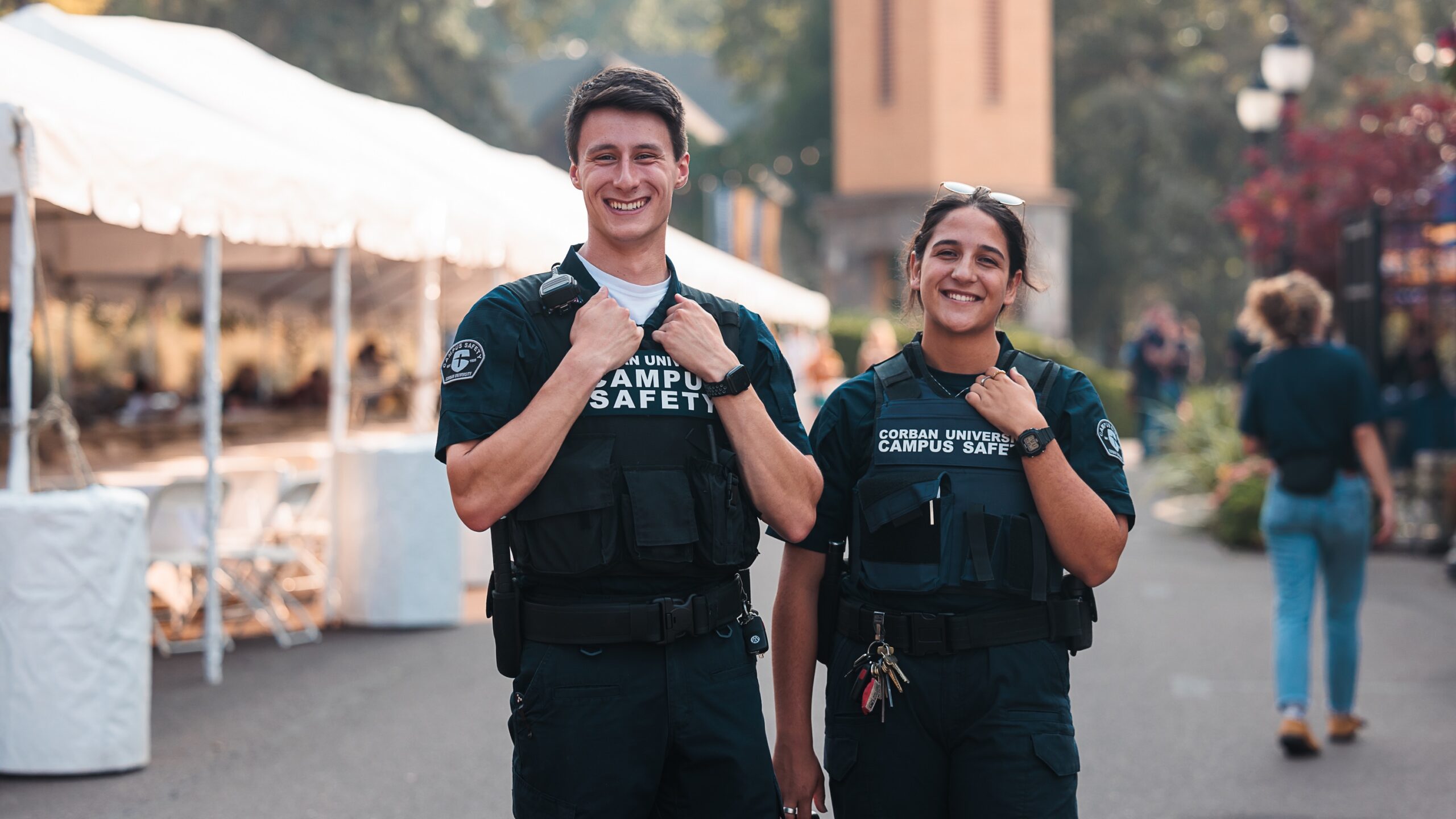 Two campus safety officers are positioned in front of tents, overseeing the surroundings for safety.