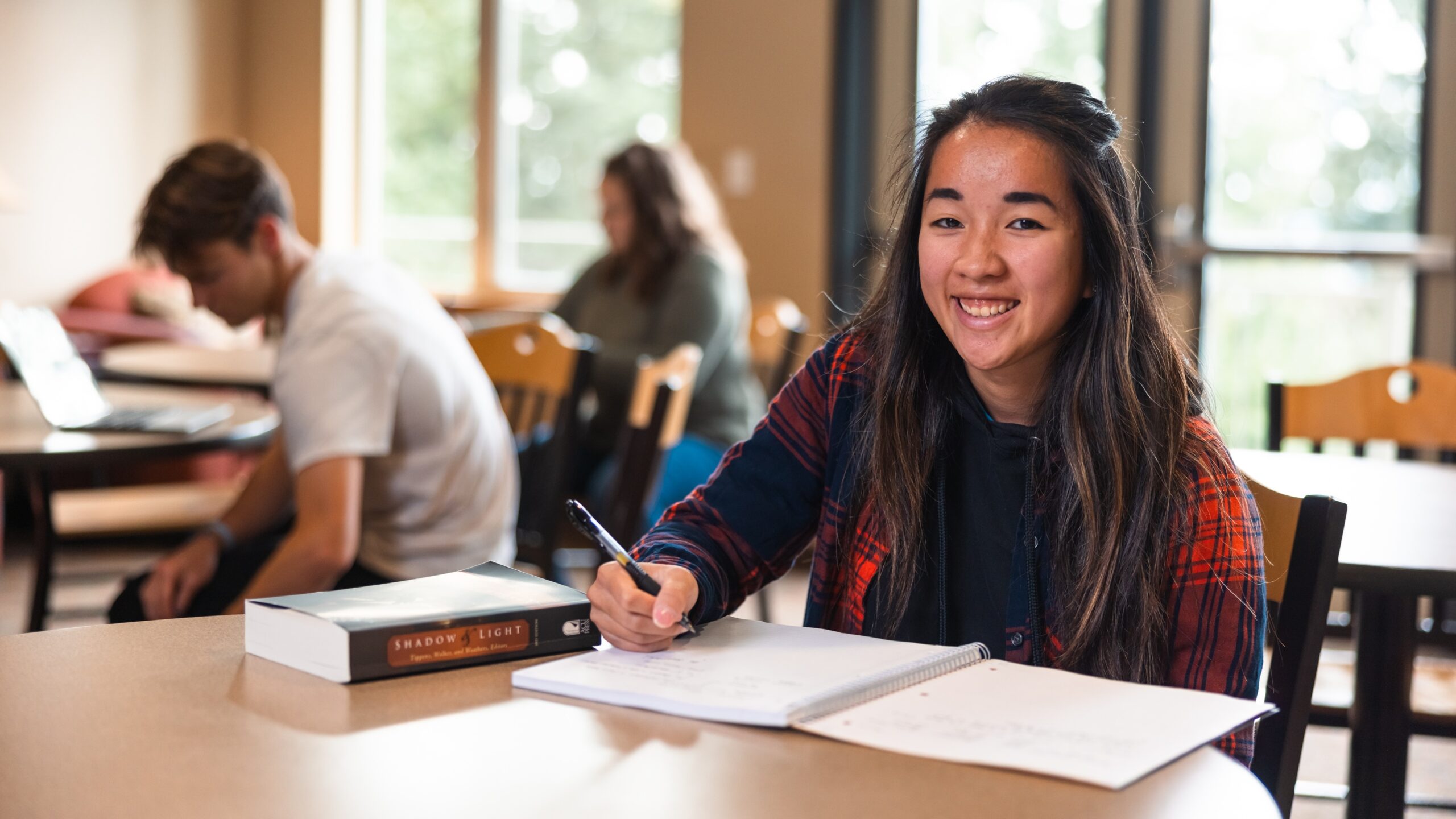 A young woman with a bright smile sits at a table, writing in her notebook, radiating positivity.