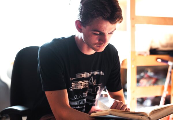 A young man is seated in a chair in a dorm room, focused on reading the Bible.
