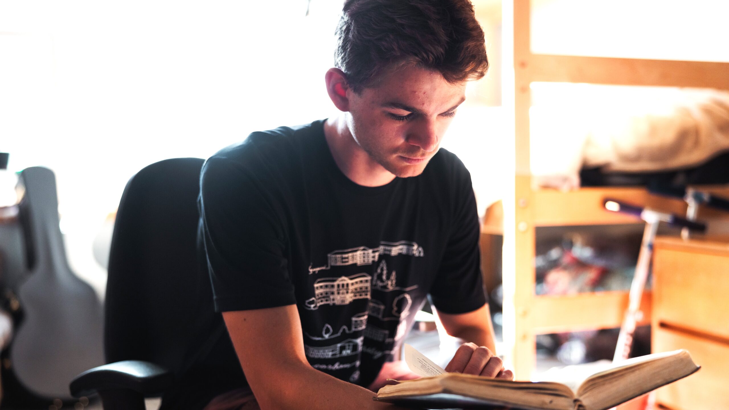 A young man is seated in a chair in a dorm room, focused on reading the Bible.