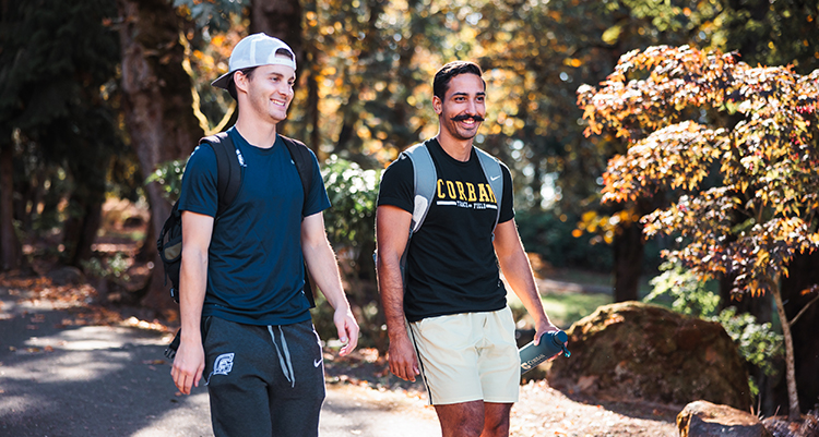 Two young men walk on a tree-lined path, smiling and carrying backpacks. One wears a backward cap, navy shirt, and sweats; the other wears a CORBAN t-shirt, light shorts, and holds a water bottle.