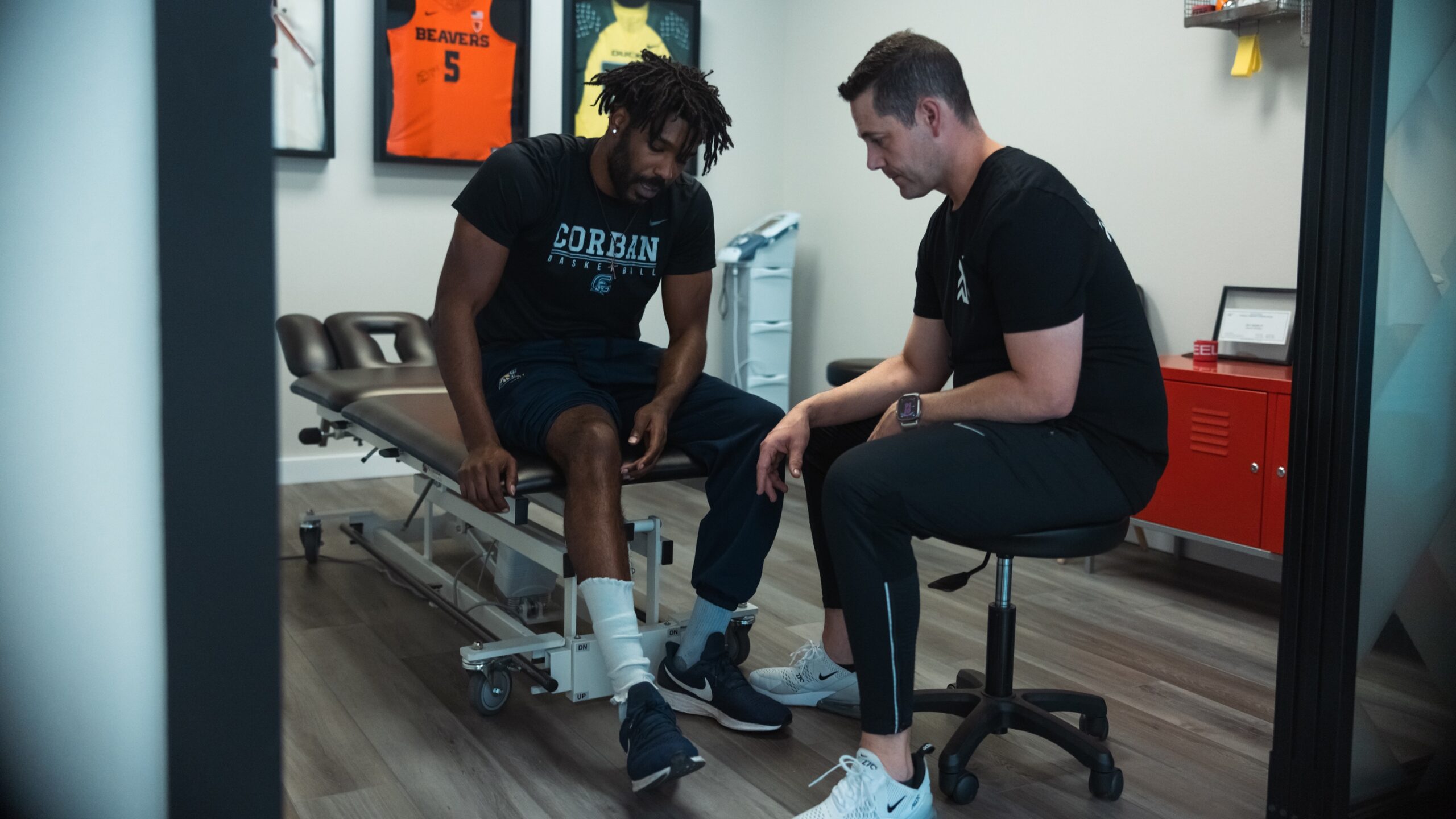 In a physical therapy room, a man sits on a bench while another man stands beside him, discussing treatment.