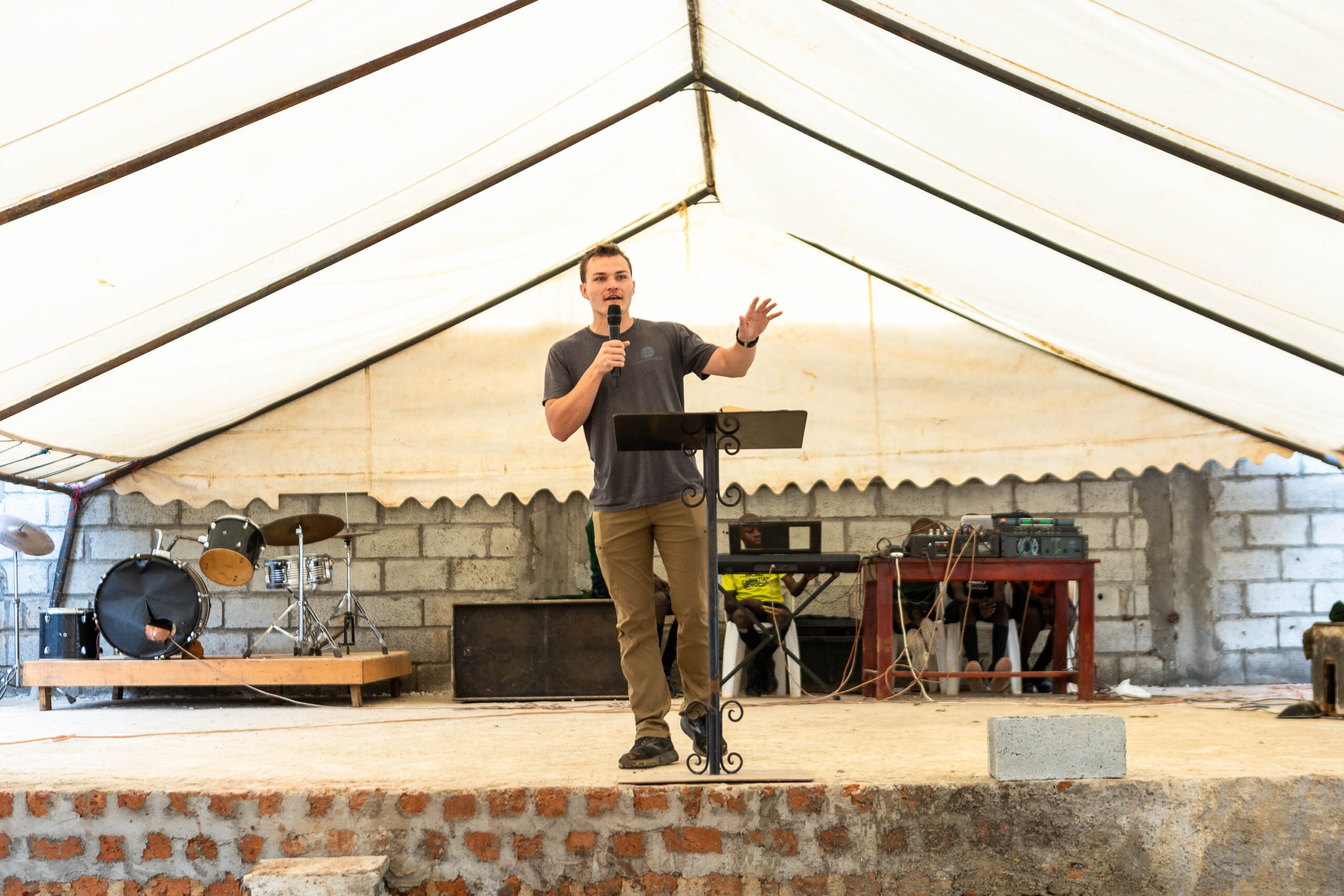 A man in Uganda stands on a stage under a large tent, addressing an audience with a microphone in hand.